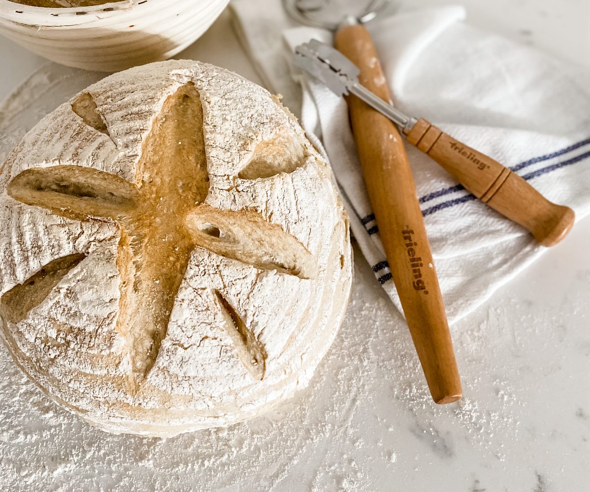 A scored sourdough loaf beside a Brotform and lame